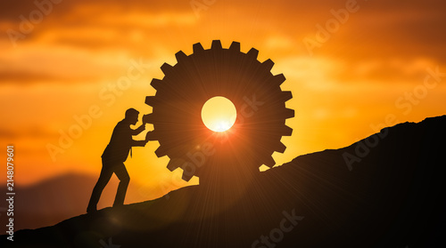 Foto Determined businessman silhouette manually pushing giant heavy gear wheel up ste
