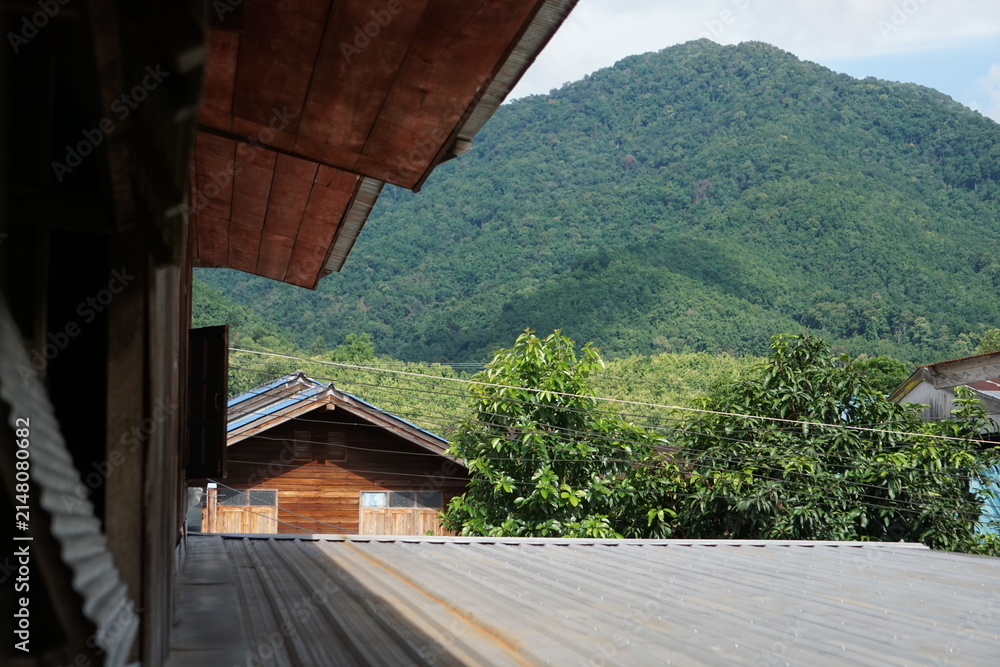 Obraz premium elevated perspective framed by traditional wooden house eaves, looking out across corrugated metal rooftops toward a massive, vibrant green mountain. The composition highlights peaceful countryside li