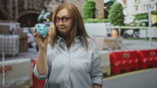 Картината върху платно Senior woman holds blue piggy bank in right hand on street with construction barriers; retirement saving confidence