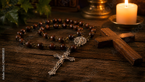 A wooden cross and a catholic rosary with a crucifix on a rustic wooden table with a candle