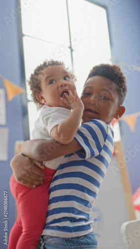 Cuadro en lienzo Older brother holds baby sister in his arms by a tall window and colorful bunting in a playroom building; affection