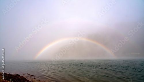 Double rainbow arching over a calm misty ocean on a foggy day with gentle waves and a rocky shore