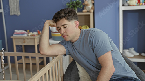 Fotografie Man seated with forearm draped over wooden crib rail, eyes closed, leaning back in a blue nursery building; quiet exhaustion parenting struggle