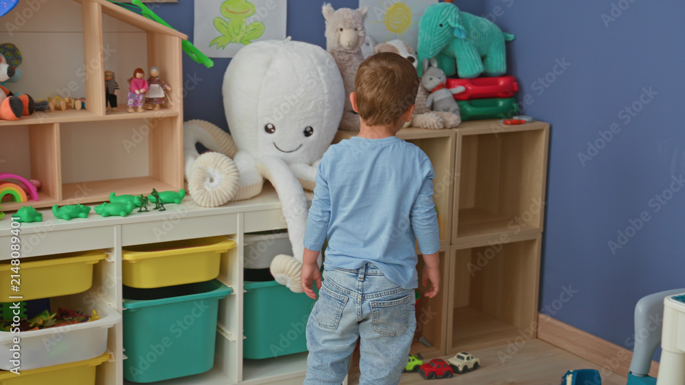 Fototapeta premium Boy picking up a yellow bucket next to a large plush octopus on shelves in a building playroom; curiosity play.