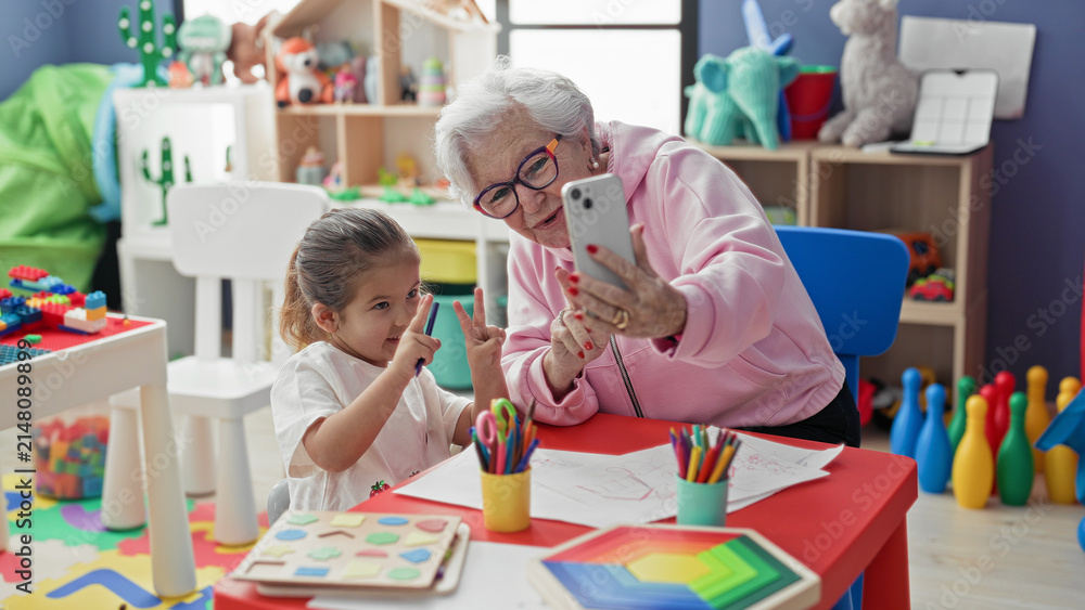 Fototapeta premium Woman holding smartphone taking selfie with little girl at colorful preschool classroom table showing drawings and colored pencils and toys on shelves; generational bonding joy.