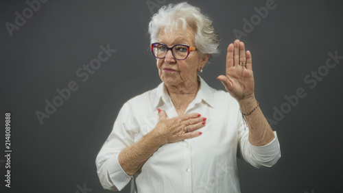 Cuadro en lienzo Elderly woman with glasses places hand on chest and raises palm in oath gesture in studio; honor sincerity