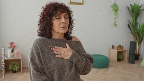 Obraz na plátně Middle age woman places hand on chest while seated with eyes closed in a yoga studio with pouf, plants and shelf visible; serenity