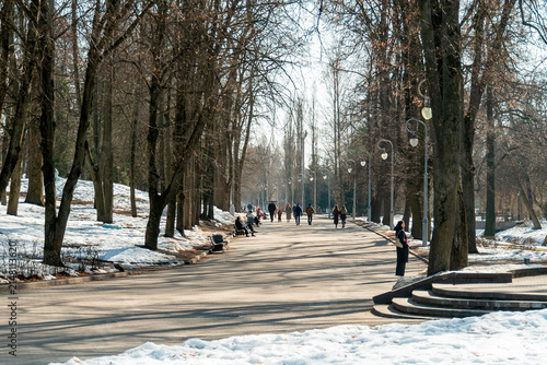 People walk along a path in a park during winter with snow under trees and light posts