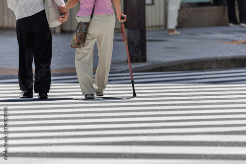 Pedestrian with a walking aid holding a companion's forearm while crossing a city street in bright daylight.
