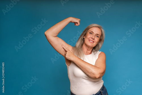 Senior woman pointing to arm flab while flexing bicep on blue background
