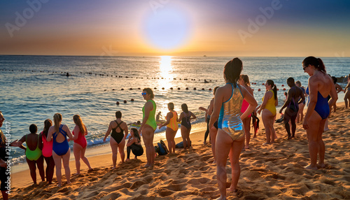 People gathering on a sandy beach during sunset, some in swimsuits.