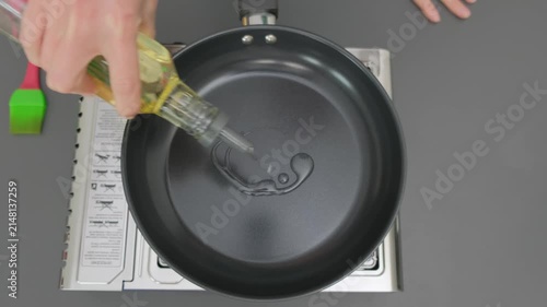 Hands pour yellow cooking oil from bottle into dark gray frying pan on gas stove in Cooking kitchen, overhead view, cooking process.