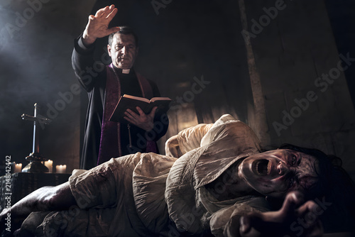 Priest performing exorcism ritual with bible over a screaming possessed person in dark candlelit room