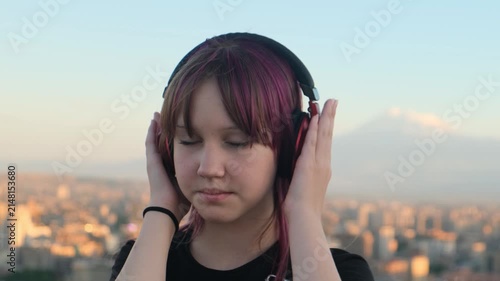 Teenage girl with purple highlighted hair listens to music in wireless headphones with Mount Ararat and Yerevan behind her. Eyes closed in peaceful enjoyment. urban lifestyle and personal wellbeing.