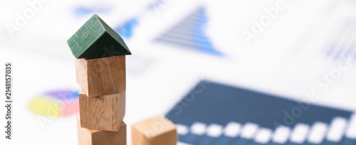 Small wooden house with green roof sits atop stacked blocks. Background holds blurred charts and graphs, hinting at data. Focus sharp on house, symbolizing stability or growth