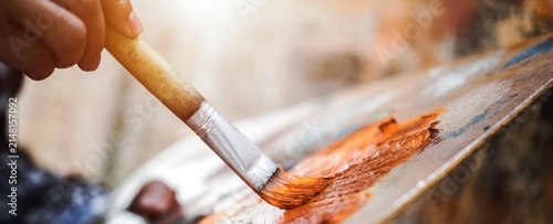 Artist's hand brushes warm paint on a palette. Sunlight glows softly through the background. Focus lies on the bristles and rich, earthy tones. Imagery evokes creativity, passion, and artistic process
