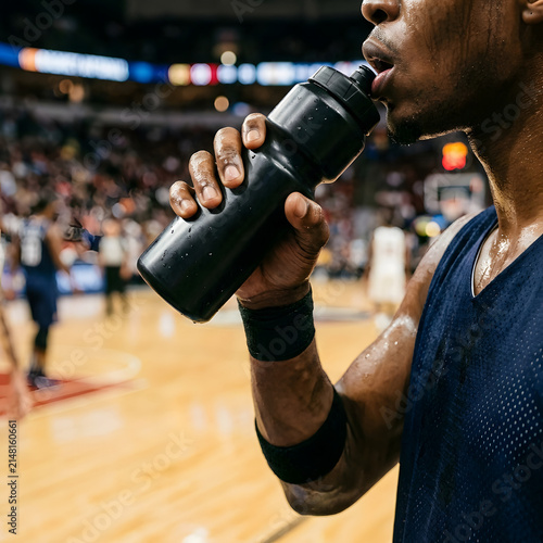 Male basketball player hydrating with a black water bottle on the court during a game or practice, showing sweat and focus.