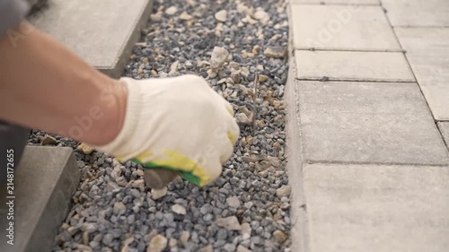 A close-up of a gloved hand laying paving slabs on a gravel base. Neutral gray tones with orange accents for safety. Landscape design, artificial turf.