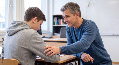 A caring father sitting next to his teenage son and putting a comforting hand on his shoulder in a classroom setting.