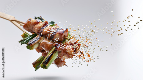 Grilled chicken skewer with glazed sauce and roasted green onion topped with flying sesame seeds, creating dynamic and appetizing food presentation