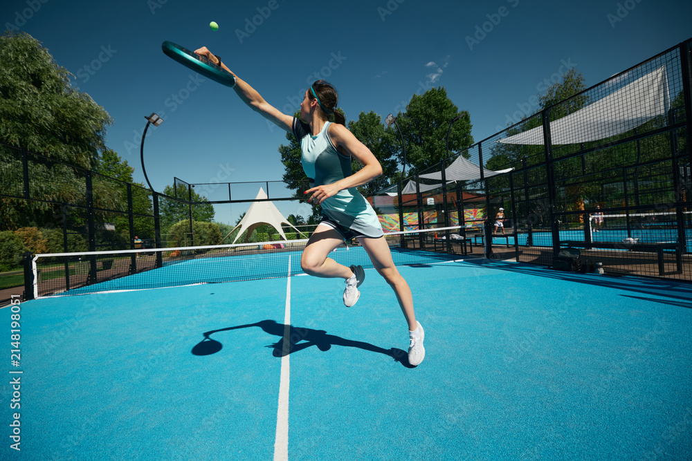 Obraz premium Stylish female padel player in blue activewear extending for high ball on sunny outdoor court. Concept of womens athletic fashion, functional sportswear, energetic brand campaign.
