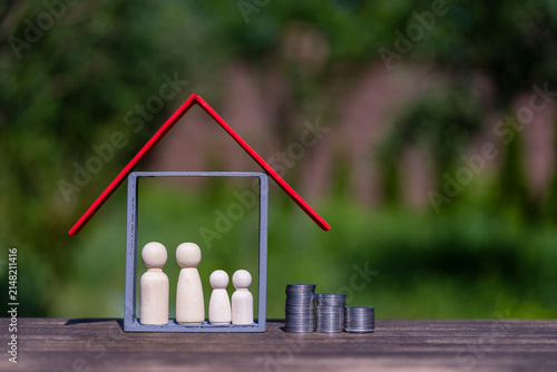Family budget and real estate mortgage investment concept. Wooden peg doll family inside house frame and stacked coins against green yard backdrop, representing property savings finance and insurance