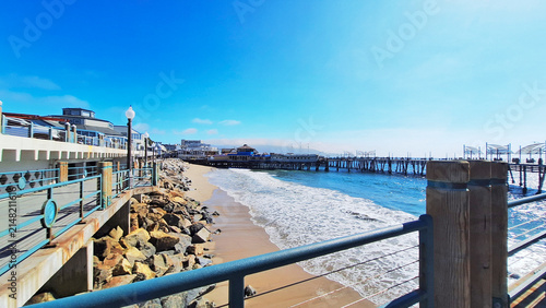 View of the historic Redondo Beach Pier structure extending into the Pacific Ocean, featuring a concrete boardwalk with railings, rocks, and ocean waves crashing on the shore. California, USA.