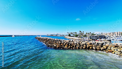 Wide shot of the stone breakwater jetty protecting King Harbor in Redondo Beach, California, showcasing clear turquoise ocean water, palm trees, and boats under a bright blue sky. USA