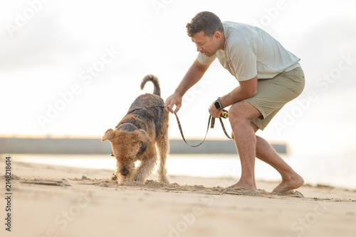 Man kneeling on sandy beach with a dog on a leash, engaging with the pet while the sun sets over the water in the background. Airedale Terrier