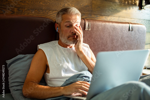 Tired middle-aged man working on laptop in bed at home