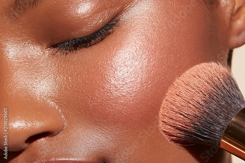 Close-up macro shot of bronzer application on cheekbone of a woman with dark skin tone, highlighting makeup brush and skin texture in a beauty setting