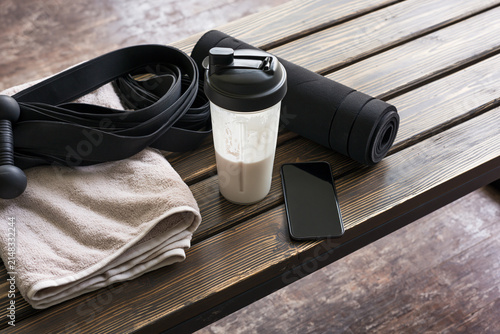 Gym equipment laid out on a wooden surface, including a shaker bottle, yoga mat, dumbbell, resistance band, towel, and smartphone. Ready for a workout