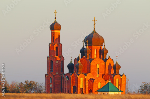 Obraz na plátně A magnificent red-brick Orthodox cathedral with dark domes illuminated by warm golden sunset light in a field