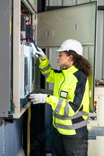 Adult female technician checks electrical control cabinet with flashlight in manufacturing workshop, inspecting wiring, control equipment and automation system during preventive maintenance.