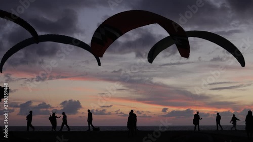 Silhouettes of paragliders and people against a vibrant sunset sky over the sea.