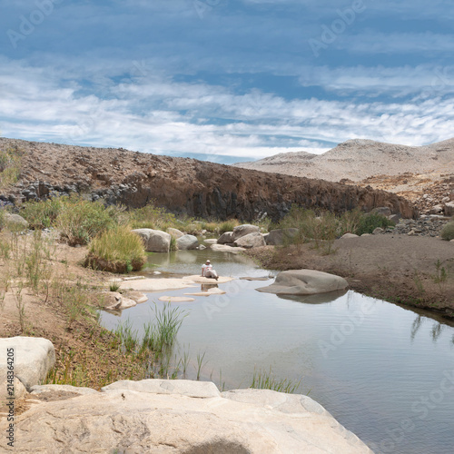 A wide landscape shot in the Afilal site captures a tranquil oasis in the Hoggar Mountains near Tamanrasset, Algeria, where a blurred person sits resting on smooth rocks beside a calm body of water.