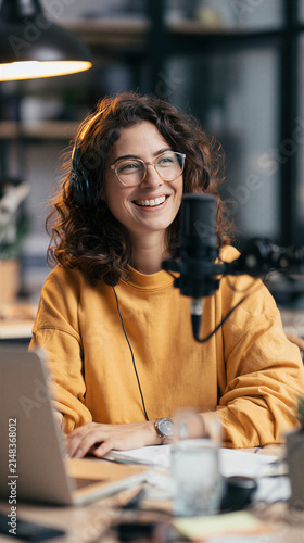 Happy young woman smiling, wearing headphones and eyeglasses, creating content, recording a podcast, doing a broadcast, or livestreaming from a home studio or office desk