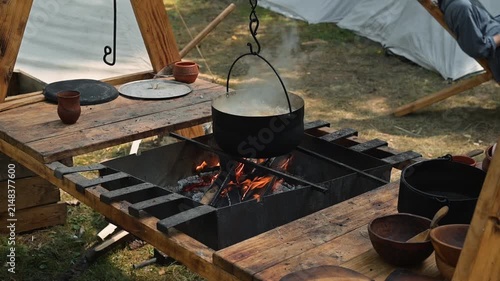 A cauldron hangs above an open fire, with flames visible in a medieval camp setting. Wooden structures and clay bowls surround the fire, highlighting daily life in the early Middle Ages