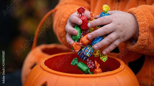 Child hand Halloween candy pumpkin bucket trick or treat orange costume Close up child hand dropping wrapped candy into pumpkin bucket during Halloween trick or treat, bright sweets, autumn