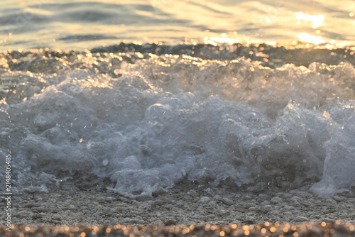  Sea wave with white foam crashing on pebble beach under golden sunset light close up background