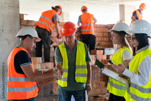 Foreman points at brick in coworker hands during materials check. Team observes stack and notes quantity while supervisor confirms delivery