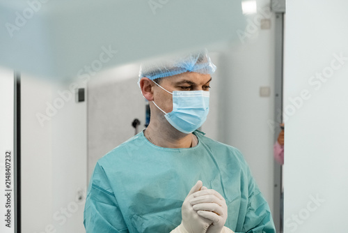 Man surgeon stands in sterile corridor with mask and gloves before operation. Focused medic clasps hands preparing for surgery behind glass