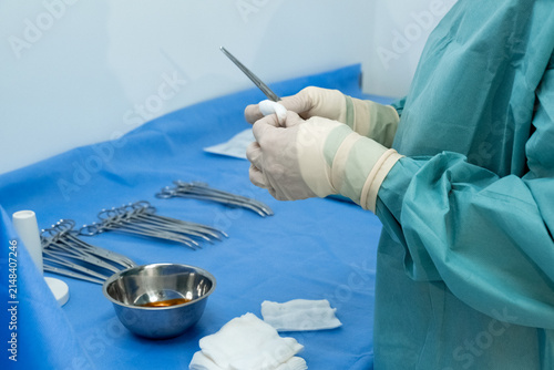 Surgeon holds metal forceps above blue sterile drape in operating room. Gloved medic checks instrument tip preparing tool for surgery