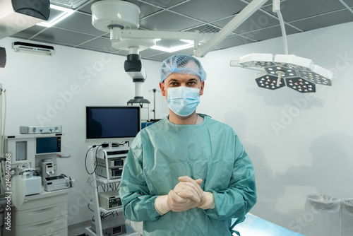 Man surgeon stands centered in operating room with mask and gloves. Focused medic clasps hands waiting calmly under bright surgical lights