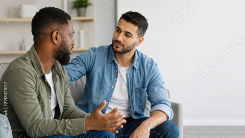 Two men engage in a supportive conversation in a bright, modern living room, with one man offering comfort and reassurance to the other.