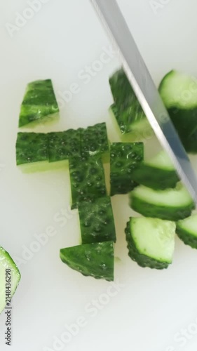 Fresh cucumber cutting close up on white kitchen board. Chef hands in blue gloves mince green vegetable with big iron knife. Cooking, preparing ingredients in professional restaurant. Top down view