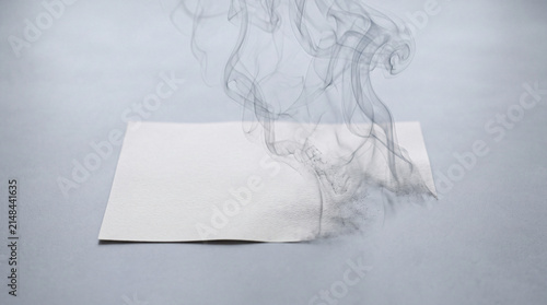 Single sheet of paper smolders at one corner, emitting curling smoke and scattered ash on a neutral gray surface, evoking themes of destruction, erasure, secrecy and slow transformation.