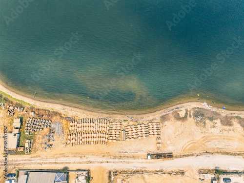 Aerial view of Kali Baru showing rows of stacked wooden materials on the sandy shore next to the calm sea, with nearby industrial structures and a coastal road in Cilincing, Jakarta, Indonesia.