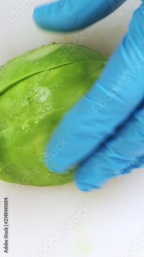 Ripe avocado cutting close up on white kitchen board. Chef hands in blue gloves mince fresh vegetable with big iron knife. Cooking, preparing ingredients in professional restaurant. Top down view