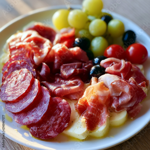 Illustration: A summer cold meat platter featuring thin slices of Italian salami, prosciutto di Parma, bresaola, and pancetta, served with soft cheese, green grapes, cherry tomatoes, and black olives.